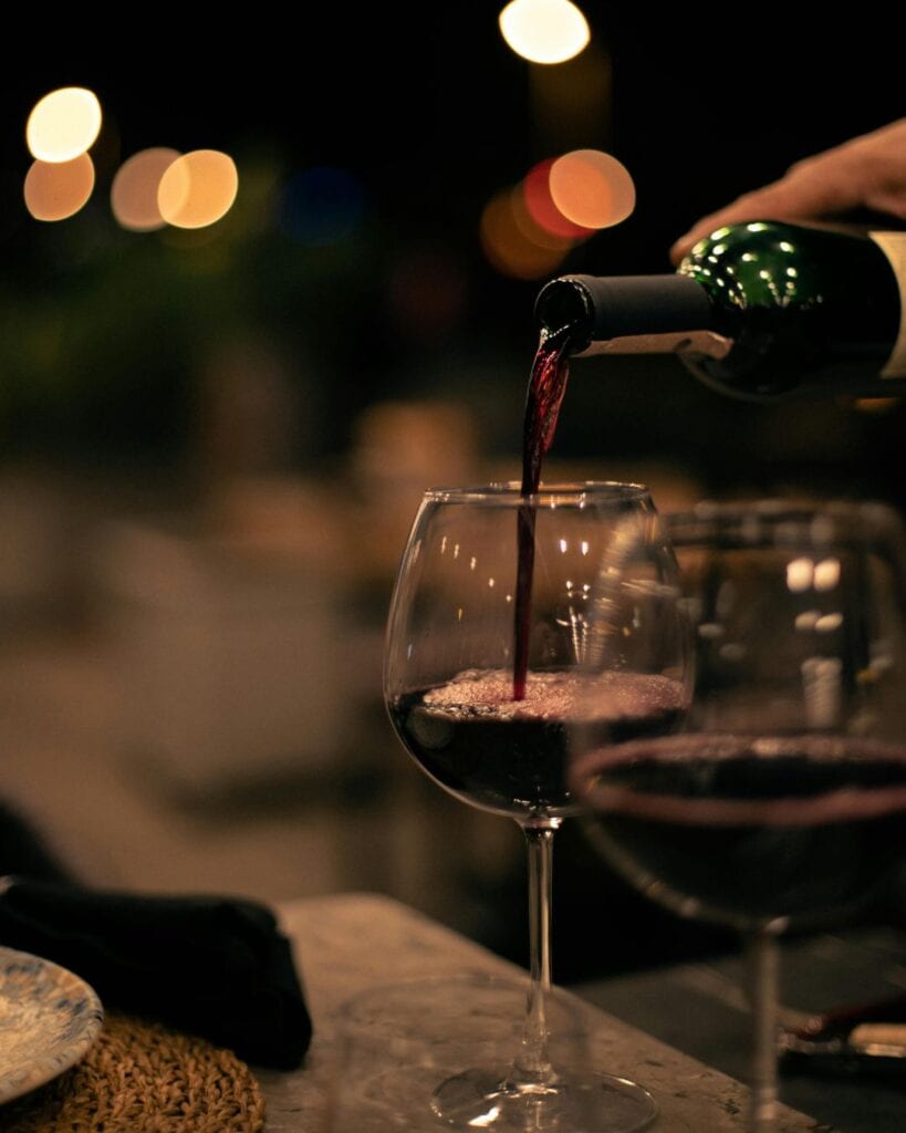 Rotwein wird am Abend aus einer Weinflasche in ein elegantes Weinglas eingeschenkt. Im stimmungsvoll verschwommenen Hintergrund leuchten herbstliche Lichter (Bokeh-Effekt). Präsentiert von bona-mente.ch für den Basler Wymärt Herbst.