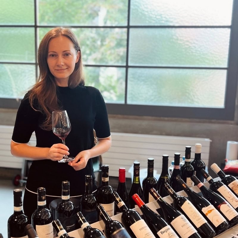 Eine Frau in einem schwarzen Oberteil hält ein Glas Rotwein in der Hand und lächelt in die Kamera. Vor ihr steht eine grosse Auswahl an verschiedenen Weinflaschen auf Tischen, im Hintergrund ist ein grosses Sprossenfenster zu sehen.