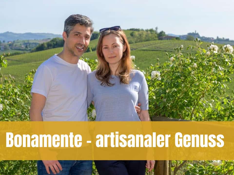 Ein lächelndes Paar steht Arm in Arm vor einer grünen Weinberg-Landschaft unter blauem Himmel. Im Vordergrund sind weisse Rosenbüsche zu sehen, und über dem Bild liegt ein gelber Banner mit dem Text „Bonamente - artisanaler Genuss“.