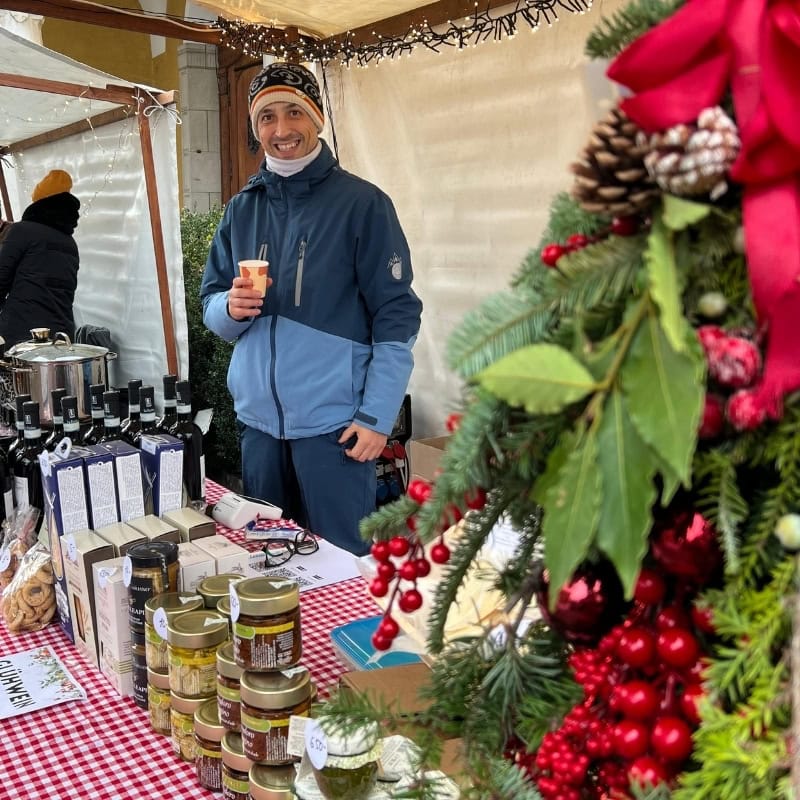 Ein Verkäufer lächelt am Stand am Gerliswiler Weihnachtsmarkt in Emmen und hält einen Becher Glühwein, im Vordergrund kunstvolle Adventsdeko mit Tannenzweigen und roten Beeren